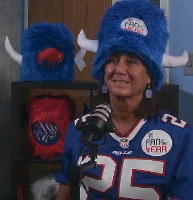 Person wearing a blue Buffalo Bills jersey and Water Buffalo Club hat with a microphone, with decorative items in the background.