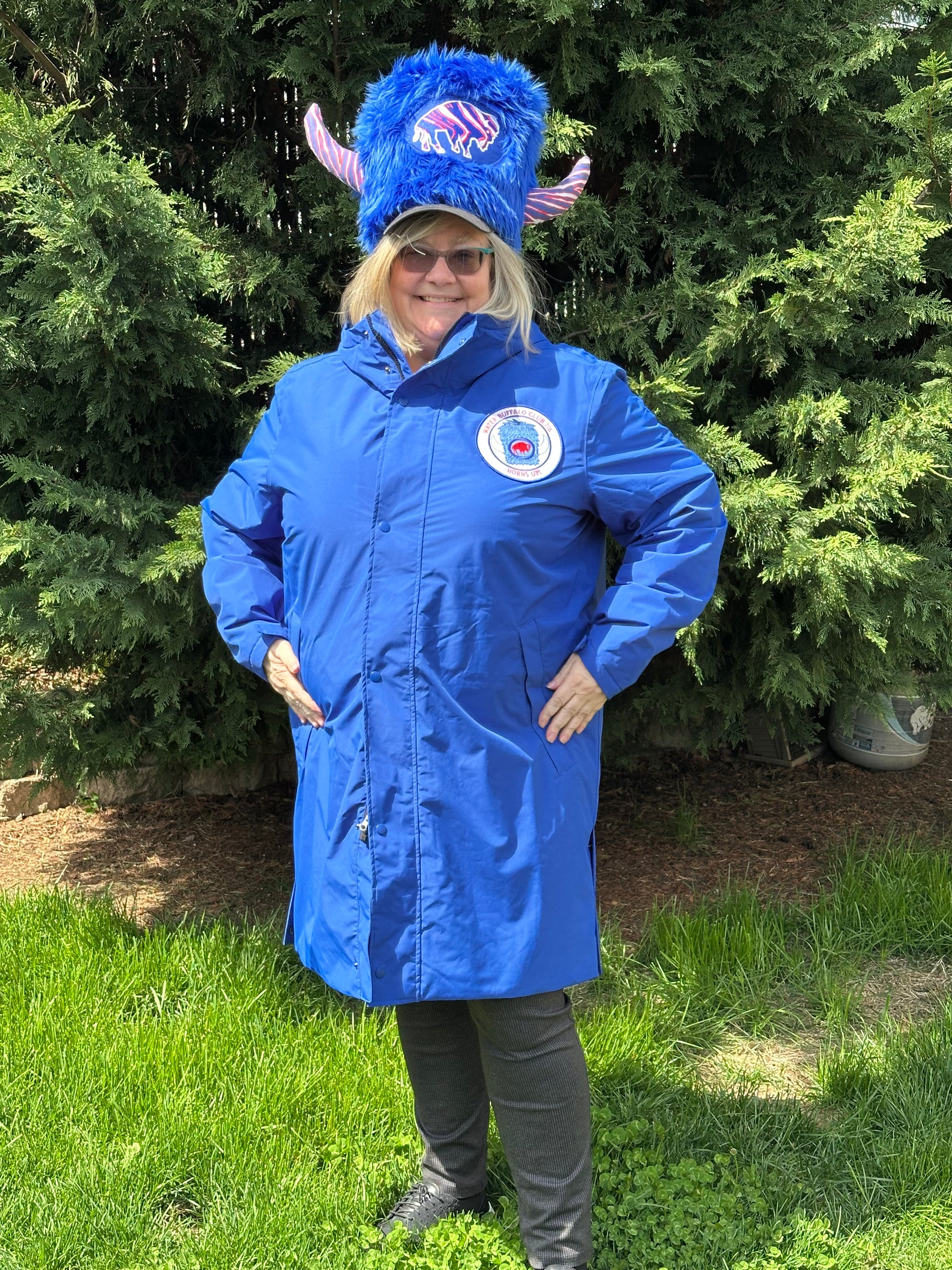 A person wearing a long royal blue stadium jacket with a Water Buffalo signature logo on the left chest panel and a Water Buffalo Club charm as a zipper pull. They are also wearing a hat with a similar logo.